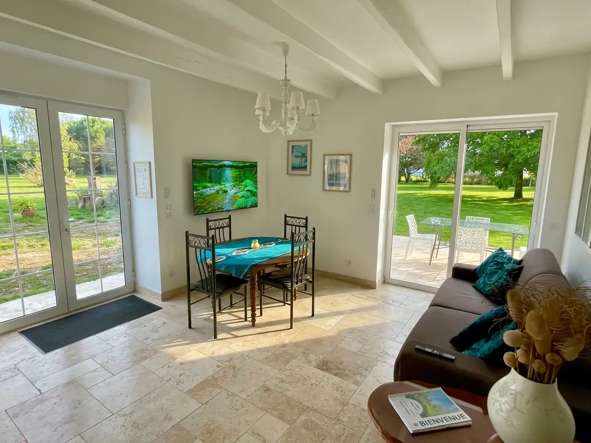 Pièce à vivre lumineuse avec sol en travertin du gîte pour curistes au Domaine des Ânes, comprenant une table avec nappe bleue, un salon avec canapé, et deux grandes portes-fenêtres donnant sur la terrasse et le jardin arboré à Tugéras-Saint-Maurice