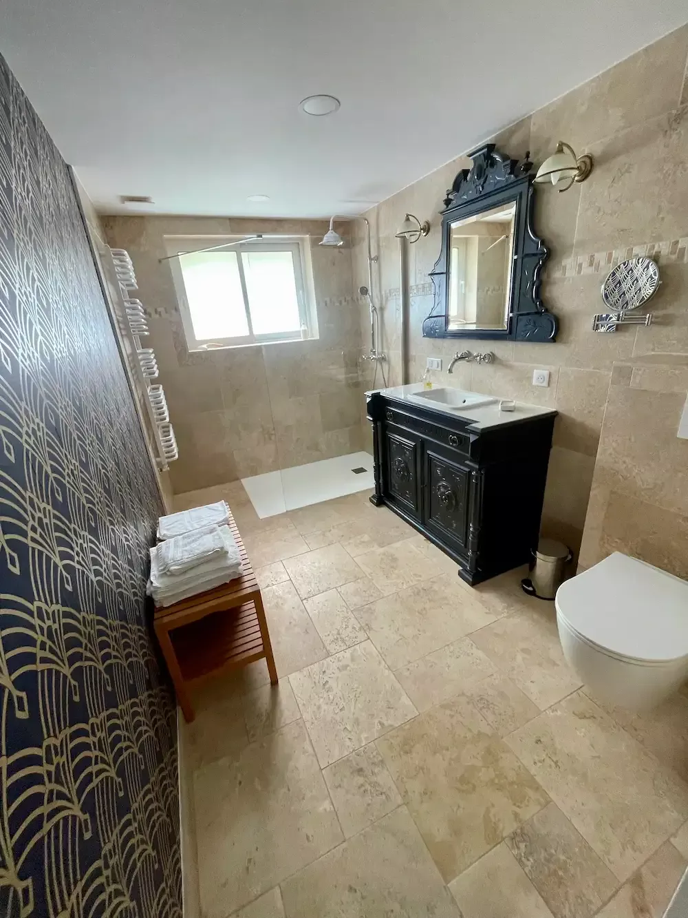 Grande salle de bain avec sol et murs en travertin du gîte pour curistes au Domaine des Ânes, comprenant une douche à l'italienne de plain-pied, un meuble vasque noir de style ancien avec miroir, un radiateur sèche-serviettes et des WC suspendus à Tugéras-Saint-Maurice à 10 min des thermes de Jonzac